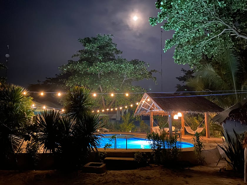 Piscina il·luminada a Malapascua de nit, amb llumets decoratius i la lluna brillant al cel.